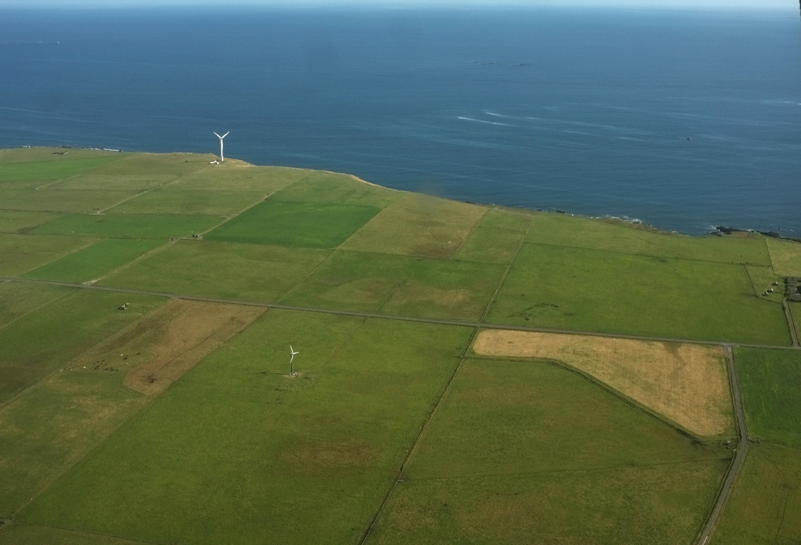 Two wind turbines standing in green coastal fields beside the sea.